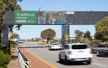 Cars drive along a multi-lane road beneath an overhead pedestrian bridge in a suburban area. The bridge features a large green advertisement reading “Do more you in Karrinyup” alongside smaller text “
